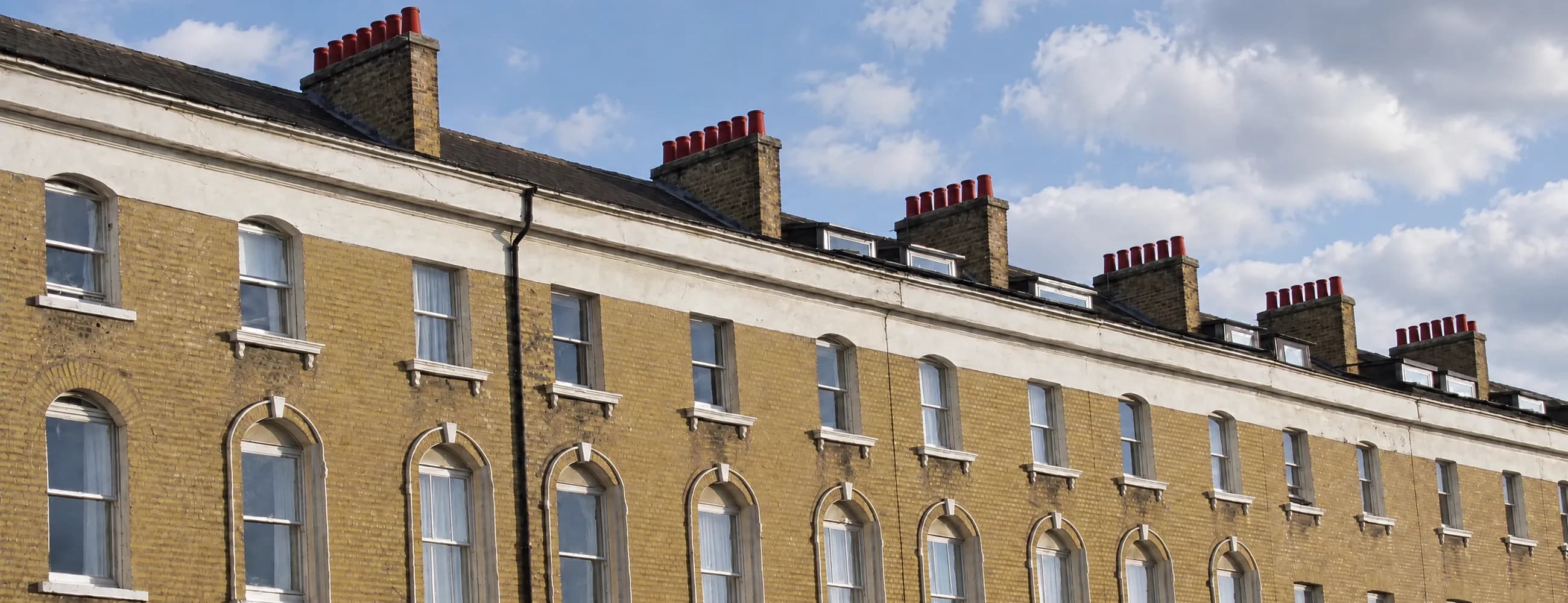 A long Victorian terrace of yellow stock brick with white sash windows, arched ground floor windows and chimney pots under a blue sky with drifting cloud
