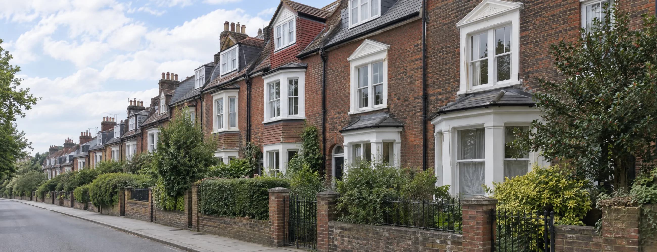 A leafy street of tall Victorian brick houses with white window surrounds, bay windows and dormers behind low brick walls, railings and hedges