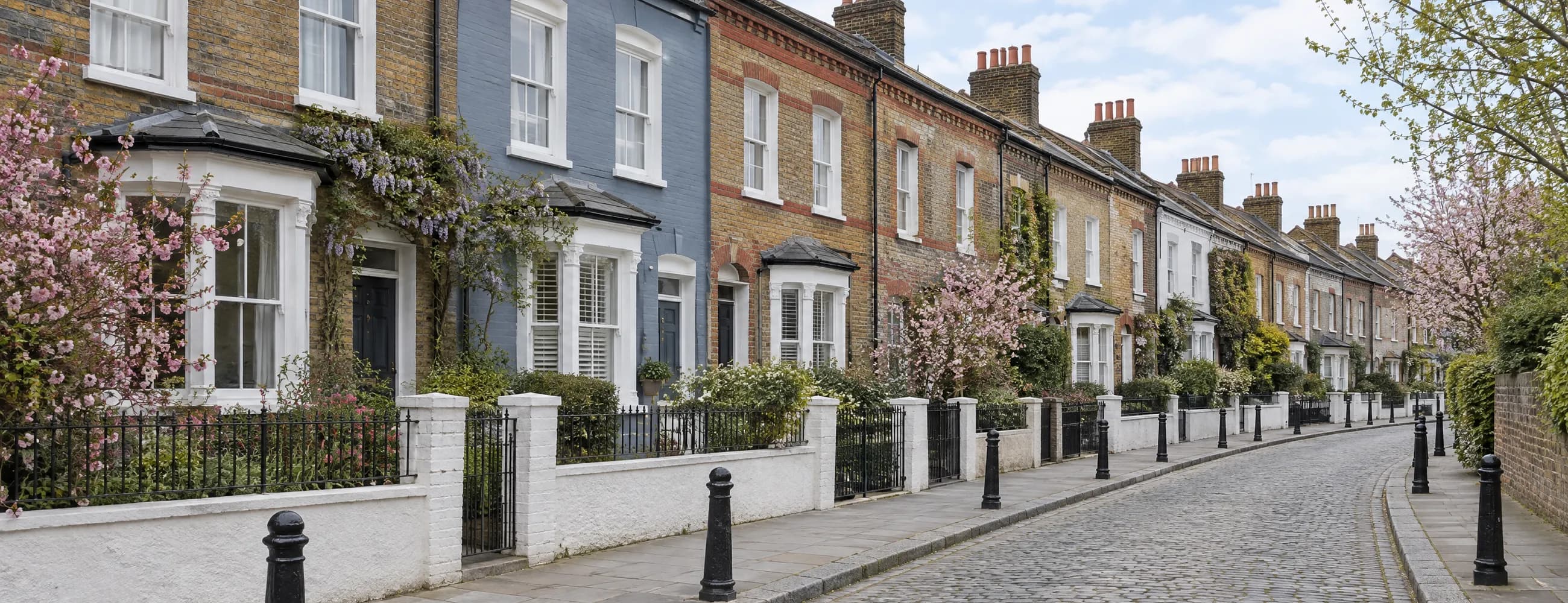 A curving cobbled side street of Victorian terraced houses in stock brick, one painted blue, behind white garden walls, railings and spring blossom